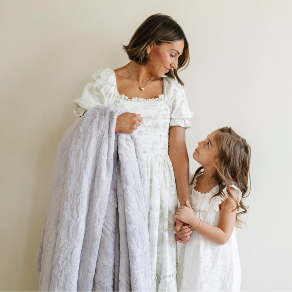 A mother stands by her daughter and holds a dove colored patterned faux fur throw Saranoni Luxury Blanket. The soft blanket is a throw blanket.