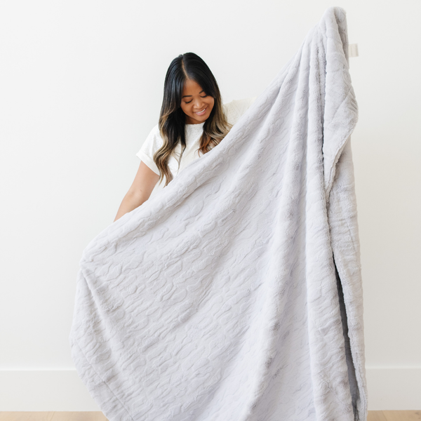 A woman smiles and holds up a light gray luxurious patterned faux fur extra large throw blanket that is a Saranoni blanket. This ultra-soft, cozy blanket adds a touch of elegance and warmth to any space, perfect as a soft oversized blanket or fluffy blanket for lounging. Ideal for those seeking a high-quality artificial fur blanket or luxury blanket, this versatile and stylish soft blanket enhances both comfort and decor. Shop the best blankets and throws for ultimate relaxation and cozy vibes.