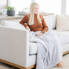 A woman sits on a couch and has a light gray luxurious patterned faux fur throw blanket that is a Saranoni blanket on her lap. This ultra-soft, plush throw blanket adds a touch of elegance and warmth to any space, perfect as a soft throw blanket or fluffy blanket for lounging. Ideal for those seeking a high-quality artificial fur blanket or luxury blanket, this versatile and stylish soft blanket enhances both comfort and decor. Shop the best blankets and throws for ultimate relaxation and cozy vibes.