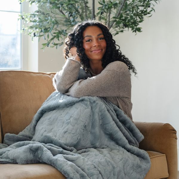 A woman sits with a dusty blue luxurious patterned faux fur throw blanket that is a Saranoni blanket. This ultra-soft, plush throw blanket adds a touch of elegance and warmth to any space, perfect as a soft throw blanket or fluffy blanket for lounging. Ideal for those seeking a high-quality artificial fur blanket or luxury blanket, this versatile and stylish soft blanket enhances both comfort and decor. Shop the best blankets and throws for ultimate relaxation and cozy vibes.