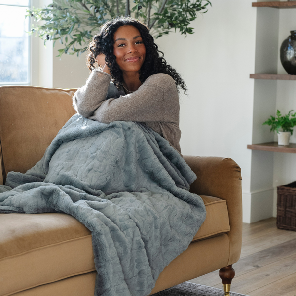 A woman smiles and sits with a dusty blue luxurious patterned faux fur extra large throw blanket that is a Saranoni blanket is on a bed. This ultra-soft, cozy blanket adds a touch of elegance and warmth to any space, perfect as a soft oversized blanket or fluffy blanket for lounging. Ideal for those seeking a high-quality artificial fur blanket or luxury blanket, this versatile and stylish soft blanket enhances both comfort and decor. Shop the best blankets and throws for ultimate relaxation and cozy vibes.