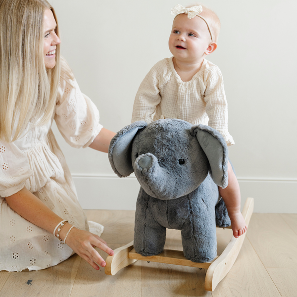 A happy little girl toddler sits on a soft gray plush elephant rocker, beaming with joy while interacting with a smiling adult beside her. The child is dressed in a cream-colored outfit and a cute hair bow, creating a heartwarming family moment in a bright and cozy room. This delightful scene captures the essence of nurturing playtime and makes for an ideal gift idea for young children.