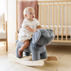 A delighted toddler in a white dress sits on a soft gray plush elephant rocker, holding onto its ears with excitement. The child has a curious expression, surrounded by a cozy nursery setting that includes a wooden crib and soft furnishings. This charming scene captures the joy of early childhood play and makes a perfect holiday gift idea for young children.