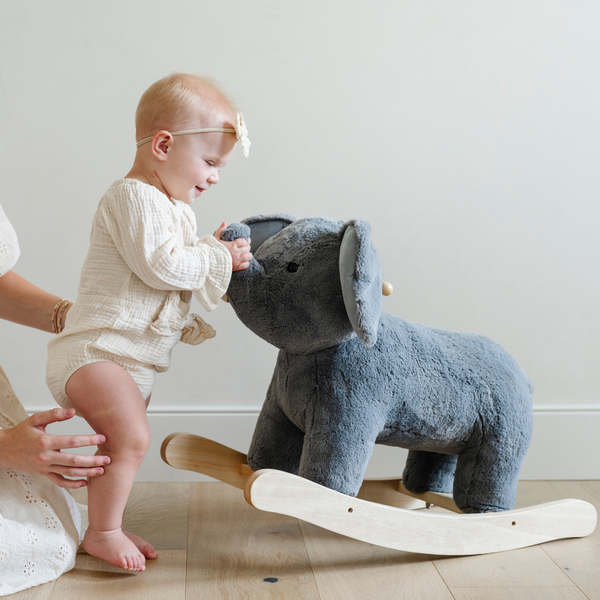 A joyful little girl toddler in a cream-colored outfit laughs as she interacts with a soft gray plush elephant rocker, reaching out to touch its trunk. A caring adult's hand is gently supporting the child, creating a heartwarming moment in a bright and cozy room. This charming scene captures the joy of early childhood play and the importance of nurturing connections, making it an ideal holiday gift idea for young children.