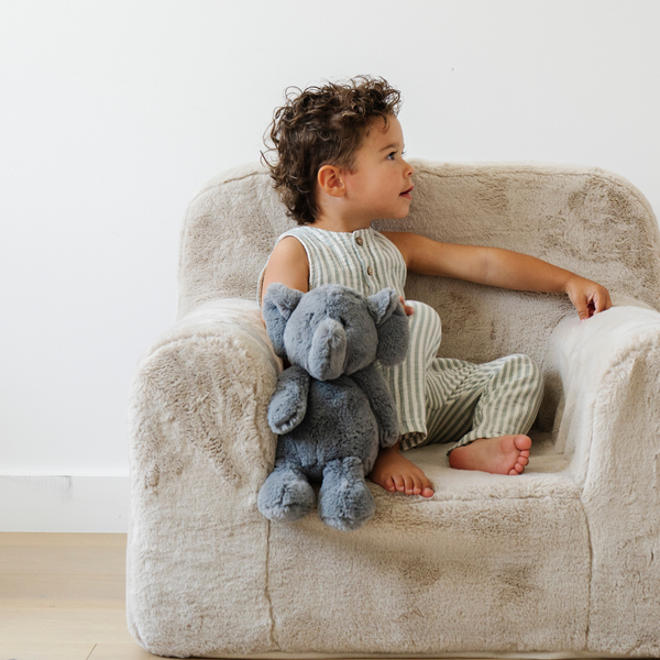 A cheerful child sits comfortably in a plush chair, holding a soft gray elephant stuffed animal. Dressed in a stylish striped outfit, the toddler gazes off to the side. This adorable scene captures the essence of childhood comfort and the joy of cuddling with plush animals, perfect for imaginative play and nurturing moments.