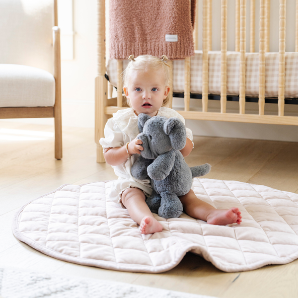 A curious toddler sits on a soft quilt, hugging a gray elephant stuffed animal. Dressed in a cute outfit, the child gazes at the camera, surrounded by a cozy nursery setting with a crib and soft blankets in the background. This heartwarming scene captures the joy of childhood and the comfort of cuddling with plush toys, perfect for nurturing imaginative play.