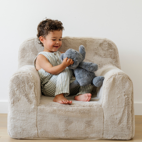 A cheerful child sits comfortably in a plush chair, holding a soft gray elephant stuffed animal. Dressed in a stylish striped outfit, the toddler gazes at the stuffed animal. This adorable scene captures the essence of childhood comfort and the joy of cuddling with plush animals, perfect for imaginative play and nurturing moments.