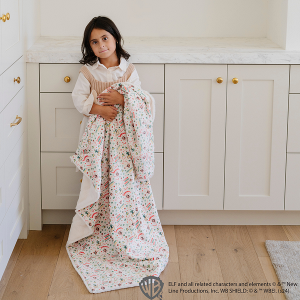 Young girl holding a cozy 'Elf' holiday blanket with a festive pattern of Buddy the Elf, candy canes, and holiday cheer messages. Ideal for holiday decor and capturing the joy of the season. The soft blanket is a luxury blanket and a Saranoni blanket. 