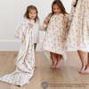 Two young girls smiling while one is wrapped in 'Elf' holiday-themed blanket with a fun pattern featuring Buddy the Elf and Christmas-themed icons and the other is wearing an 'Elf' snuggler with the same pattern. Perfect for bringing festive joy and comfort to the holiday season.