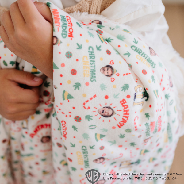 Close-up of an 'Elf' holiday-themed blanket with a festive design featuring Buddy the Elf, candy canes, and phrases like 'Christmas Cheer' and 'Cotton-Headed Ninny.' Perfect for adding fun and whimsy to holiday decor. The soft blanket is a luxury blanket and a Saranoni blanket. 