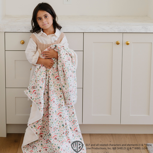 Young girl holding a cozy 'Elf' holiday blanket with a festive pattern of Buddy the Elf, candy canes, and holiday cheer messages. Ideal for holiday decor and capturing the joy of the season. The soft blanket is a luxury blanket and a Saranoni blanket. 