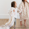 Two young girls smiling while one is wrapped in 'Elf' holiday-themed blanket with a fun pattern featuring Buddy the Elf and Christmas-themed icons and the other is wearing an 'Elf' snuggler with the same pattern. Perfect for bringing festive joy and comfort to the holiday season. The soft blanket is a luxury blanket and a Saranoni blanket. 