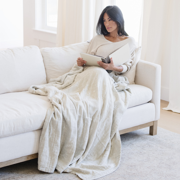 A woman sitting with a Saranoni XL muslin quilt in a soft neutral print. This luxury blanket features four layers of ultra-soft bamboo muslin, creating an airy yet cozy feel. Designed with a stitched quilted pattern and a one-inch border, this breathable blanket is perfect for muslin lovers looking for extra warmth and comfort. Made from bamboo fabric, this soft muslin quilt is a lightweight quilt that offers a cozy, neutral home décor accent. The softest blanket and a Saranoni blanket.