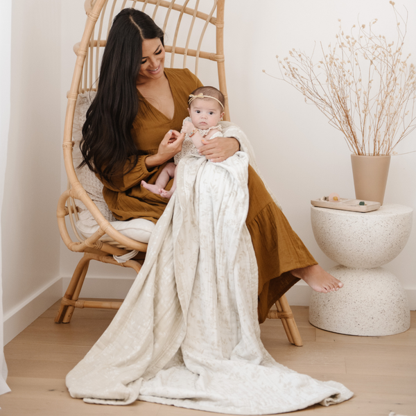 A woman and her little girl sit with a Saranoni XL muslin quilt in a soft neutral print. This luxury blanket features four layers of ultra-soft bamboo muslin, creating an airy yet cozy feel. Designed with a stitched quilted pattern and a one-inch border, this breathable blanket is perfect for muslin lovers looking for extra warmth and comfort. Made from bamboo fabric, this soft muslin quilt is a lightweight quilt that offers a cozy, home décor accent. The softest blanket and a Saranoni blanket.