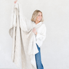 A woman holding a Saranoni XL muslin quilt in a soft neutral print. This luxury blanket features four layers of ultra-soft bamboo muslin, creating an airy yet cozy feel. Designed with a stitched quilted pattern and a one-inch border, this breathable blanket is perfect for muslin lovers looking for extra warmth and comfort. Made from bamboo fabric, this soft muslin quilt is a lightweight quilt that offers a cozy, neutral home décor accent. The softest blanket and a Saranoni blanket.