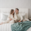 A husband and wife smile and sit by a eucalyptus luxurious patterned faux fur throw blanket that is a Saranoni blanket. This ultra-soft, plush throw blanket adds a touch of elegance and warmth to any space, perfect as a soft throw blanket or fluffy blanket for lounging. Ideal for those seeking a high-quality artificial fur blanket or luxury blanket, this versatile and stylish soft blanket enhances both comfort and decor. Shop the best blankets and throws for ultimate relaxation and cozy vibes.