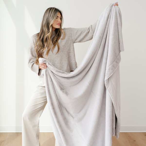 A woman holds up a soft blanket that is a light gray cozy blanket. This luxury blanket is also an extra large blanket and a Saranoni blanket. This faux fur blanket is great for gifting, relaxing, and elevating your home as a home decor staple. 
