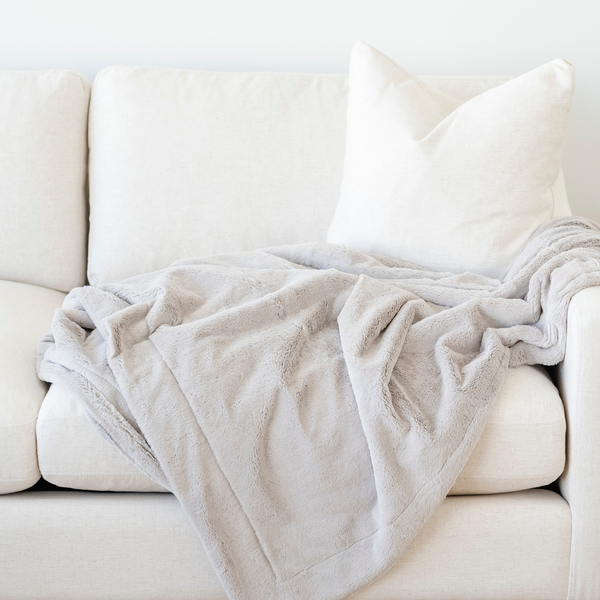 A gorgeous light gray blanket is draped on a cream couch. This soft blanket is a lush blanket and a Saranoni blanket. This luxury blanket is such a cozy blanket and is great for relaxing, gifting, or as a home decor staple. Enjoy this extra large blanket for years to come. 
