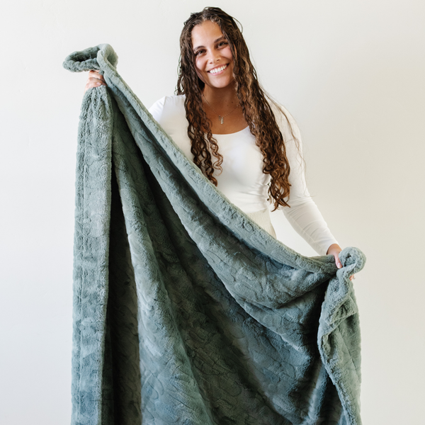 A woman smiles and holds a dusty green luxurious patterned faux fur extra large throw blanket that is a Saranoni blanket. This ultra-soft, cozy blanket adds a touch of elegance and warmth to any space, perfect as a soft oversized blanket or fluffy blanket for lounging. Ideal for those seeking a high-quality artificial fur blanket or luxury blanket, this versatile and stylish soft blanket enhances both comfort and decor. Shop the best blankets and throws for ultimate relaxation and cozy vibes.