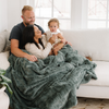 A husband, wife, and their daughter and a dusty green luxurious patterned faux fur extra large throw blanket that is a Saranoni blanket. This ultra-soft, cozy blanket adds a touch of elegance and warmth to any space, perfect as a soft oversized blanket or fluffy blanket for lounging. Ideal for those seeking a high-quality artificial fur blanket or luxury blanket, this versatile and stylish soft blanket enhances both comfort and decor. Shop the best blankets and throws for ultimate relaxation and cozy vibes.