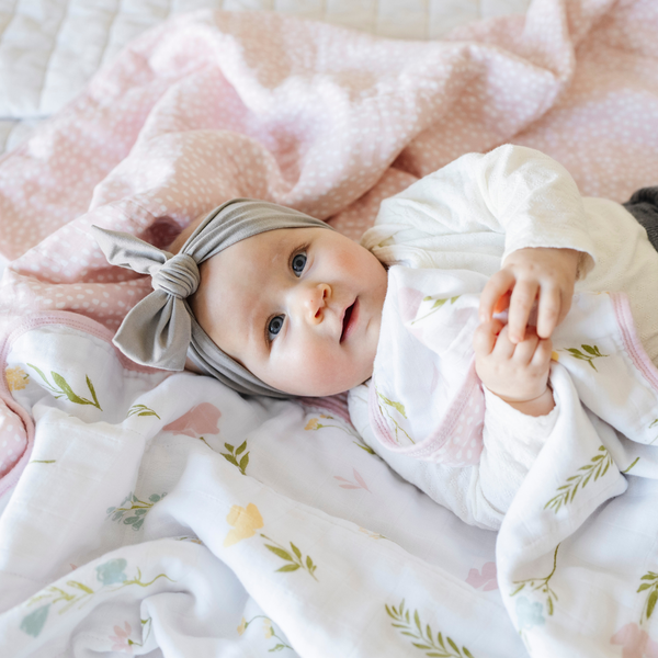 A baby girl lies on a luxuriously soft, reversible muslin quilt. The soft blanket is a luxury blanket and a lightweight and airy baby blanket as well as a Saranoni blanket. One side of the baby quilt features a delicate floral print with soft pink trim, while the reverse showcases a blush pink dotted pattern. This ultra-soft, baby blanket is perfect for snuggles, tummy time, stroller rides, or nursery decor, making it an essential for newborns, infants, and toddlers.