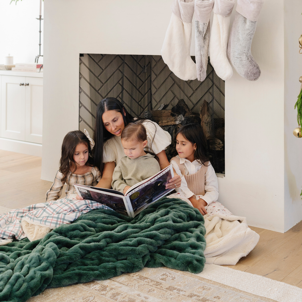 Mother reading a story to children by the fireplace, with everyone snuggled under a soft, green double ruched holiday blanket. Perfect for capturing the warmth and togetherness of family time during the holidays. The soft blanket is a luxury blanket and a Saranoni blanket. 