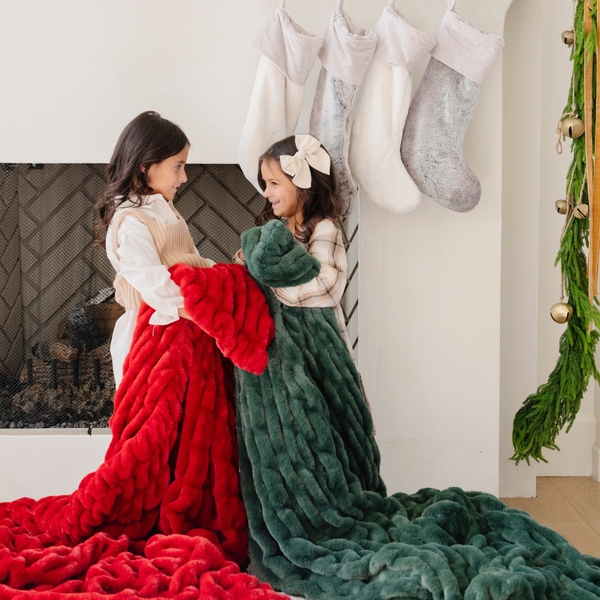 Two young girls by the fireplace with holiday stockings, each wrapped in cozy double ruched blankets—one in festive red and the other in plush green. Perfect for capturing the warmth and joy of the holiday season with luxurious, seasonal decor. The soft blankets are luxury blankets and Saranoni blankets.