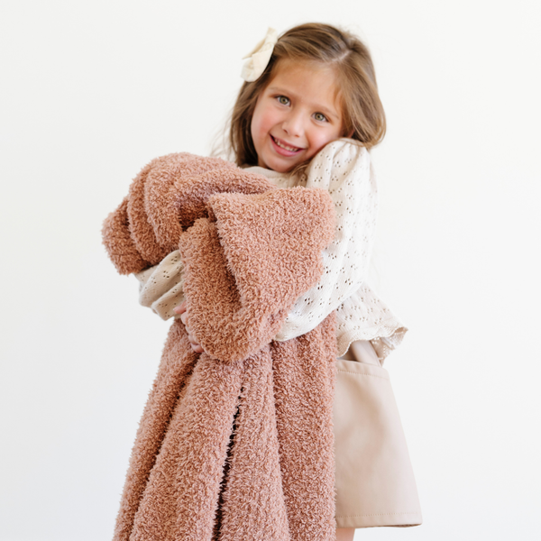 A little girl holds a Bamboni® blanket by Saranoni. This ultra-soft blanket is a clay colored blanket and made from a premium blend of bamboo rayon and poly microfiber, offering a lightweight, breathable, and fluffy feel. Perfect as a extra large blanket, luxury blanket, or soft blanket, it adds warmth and elegance to any living room, bedroom, or home décor setting. Designed for ultimate comfort, this buttery-soft Saranoni blanket is ideal for gifting. Enjoy this cozy blanket for years to come!