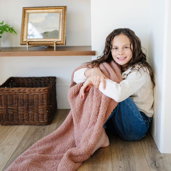 A little girl smiles and holds a Bamboni® blanket by Saranoni. This ultra-soft blanket is a clay colored blanket and made from a premium blend of bamboo rayon and poly microfiber, offering a lightweight, breathable, and fluffy feel. Perfect as a toddler blanket, luxury blanket, or soft blanket, it adds warmth and elegance to any bedroom and is a kids blanket. Designed for ultimate comfort, this buttery-soft Saranoni blanket is ideal for gifting. Enjoy this cozy blanket for years to come!