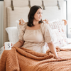 A woman sits with a luxurious ribbed Bamboni® blanket by Saranoni, the blanket is a ginger color. This ultra-soft blanket is crafted from bamboo rayon and poly microfiber, making it lightweight, breathable, and fluffy. Ideal as a cozy blanket, throw blanket, luxury blanket, soft blanket, or stylish home accent, this Saranoni blanket brings warmth, comfort, and timeless elegance to any living room or bedroom.