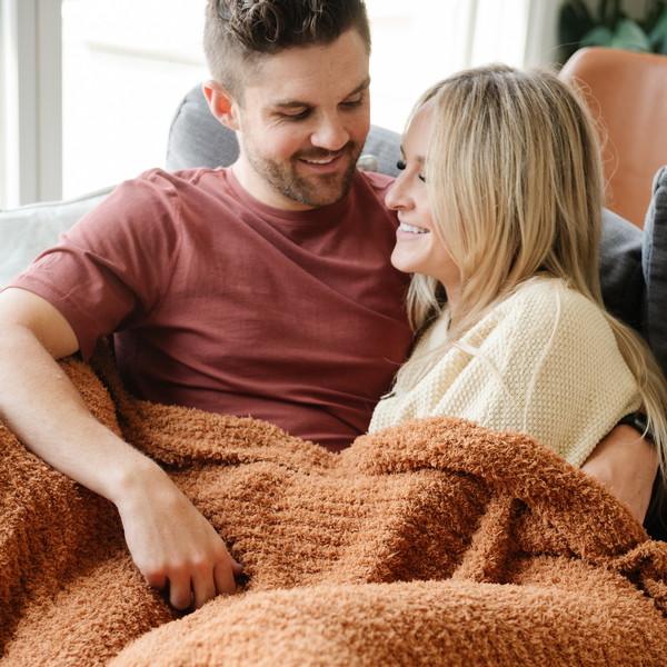 A husband and wife sit under a luxurious ribbed Bamboni® blanket by Saranoni, the blanket is a ginger color. This ultra-soft blanket is crafted from bamboo rayon and poly microfiber, making it lightweight, breathable, and fluffy. Ideal as a cozy blanket, throw blanket, luxury blanket, soft blanket, or stylish home accent, this Saranoni blanket brings warmth, comfort, and timeless elegance to any living room or bedroom.