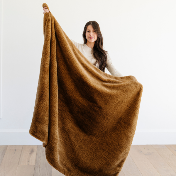 A woman holds up a Saranoni Grand Faux Fur blanket, featuring an elegant golden tone and luxuriously soft texture. This cozy, throw blanket and faux fur blanket offers soothing weight, premium softness, timeless comfort, and is a soft blanket. Perfect as a cozy blanket and luxury blanket for snuggling or as a luxury bedroom accent, this Saranoni blanket elevates every cozy moment and is a cozy blanket.