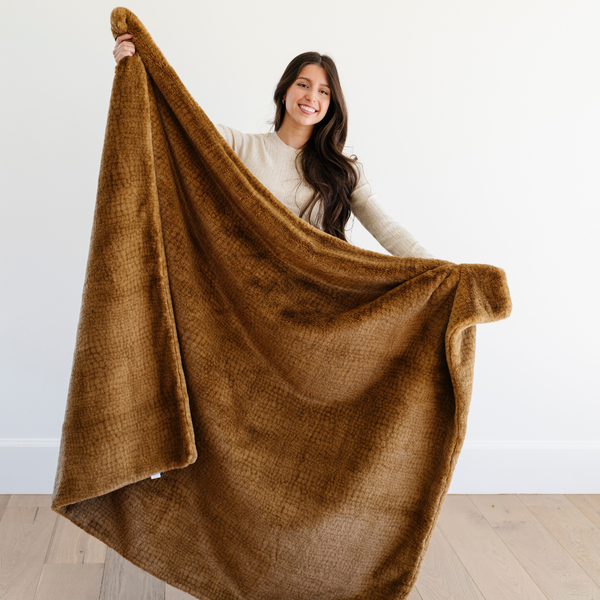 A woman smiles and holds a Saranoni Grand Faux Fur blanket, featuring an elegant golden tone and luxuriously soft texture. This cozy, throw blanket and faux fur blanket offers soothing weight, premium softness, timeless comfort, and is a soft blanket. Perfect as a cozy blanket and luxury blanket for snuggling or as a luxury bedroom accent, this Saranoni blanket elevates every cozy moment and is a cozy blanket.