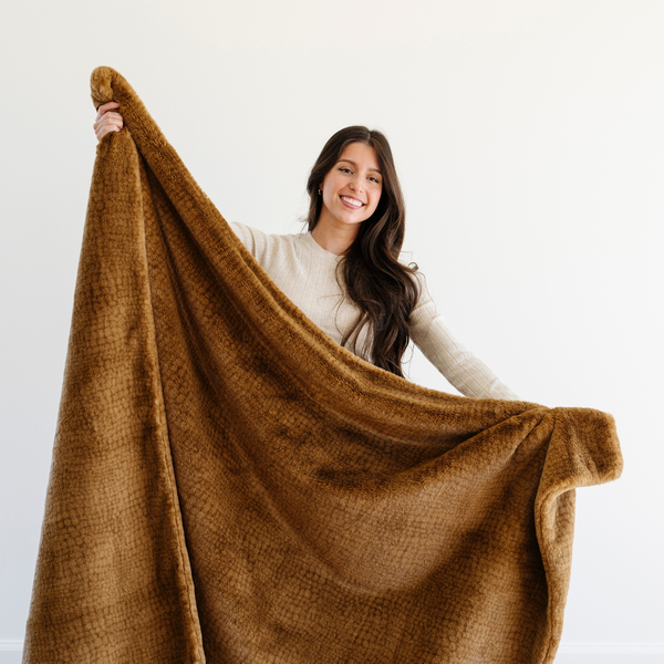 A woman smiles and holds up a Saranoni Grand Faux Fur blanket, featuring an elegant golden tone and luxuriously soft texture. This cozy, extra large throw blanket and faux fur blanket offers soothing weight, premium softness, timeless comfort, and is a soft blanket. Perfect as a cozy blanket and luxury blanket for snuggling or as a luxury bedroom accent, this Saranoni blanket elevates every cozy moment and is a cozy blanket.