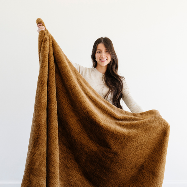 A woman holds up a Saranoni Grand Faux Fur blanket, featuring an elegant golden tone and luxuriously soft texture. This cozy, extra large throw blanket and faux fur blanket offers soothing weight, premium softness, timeless comfort, and is a soft blanket. Perfect as a cozy blanket and luxury blanket for snuggling or as a luxury bedroom accent, this Saranoni blanket elevates every cozy moment and is a cozy blanket.