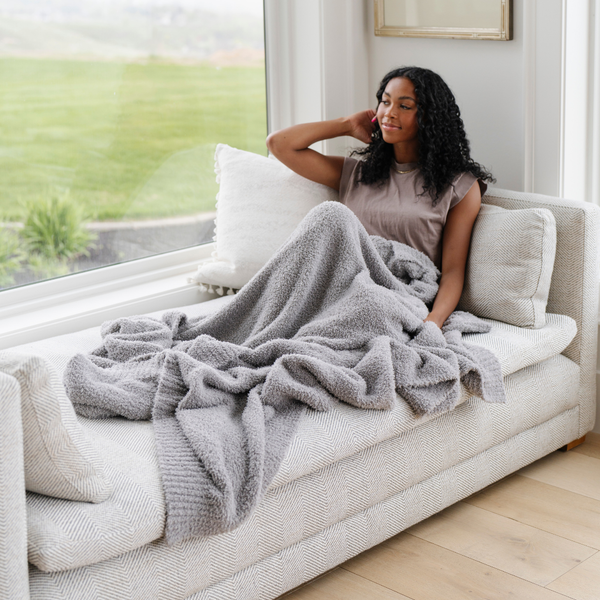 A woman sits under a Bamboni® blanket by Saranoni. This ultra-soft blanket is a gray blanket and made from a premium blend of bamboo rayon and poly microfiber, offering a lightweight, breathable, and fluffy feel. Perfect as a extra large blanket, luxury blanket, or soft blanket, it adds warmth and elegance to any living room, bedroom, or home décor setting. Designed for ultimate comfort, this buttery-soft Saranoni blanket is ideal for gifting. Enjoy this cozy blanket for years to come! 