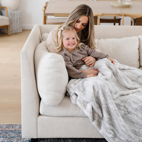 A mother and her daughter sit under a Saranoni blanket, specifically the gray marbled faux fur/minky throw, highlighting its ultra-soft texture and luxurious design. This plush throw blanket features a dual-texture blend of silky minky and cozy faux fur, creating the ultimate cozy throw blanket for home decor. Perfect luxury blanket for lounging or adding style to any space. Soft blanket, cozy blanket, plush throw—experience unmatched warmth with a Saranoni throw blanket.