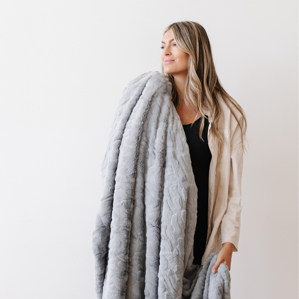 A woman smiles and holds a gray luxurious patterned faux fur extra large throw blanket that is a Saranoni blanket. This ultra-soft, cozy blanket adds a touch of elegance and warmth to any space, perfect as a soft oversized blanket or fluffy blanket for lounging. Ideal for those seeking a high-quality artificial fur blanket or luxury blanket, this versatile and stylish soft blanket enhances both comfort and decor. Shop the best blankets and throws for ultimate relaxation and cozy vibes.