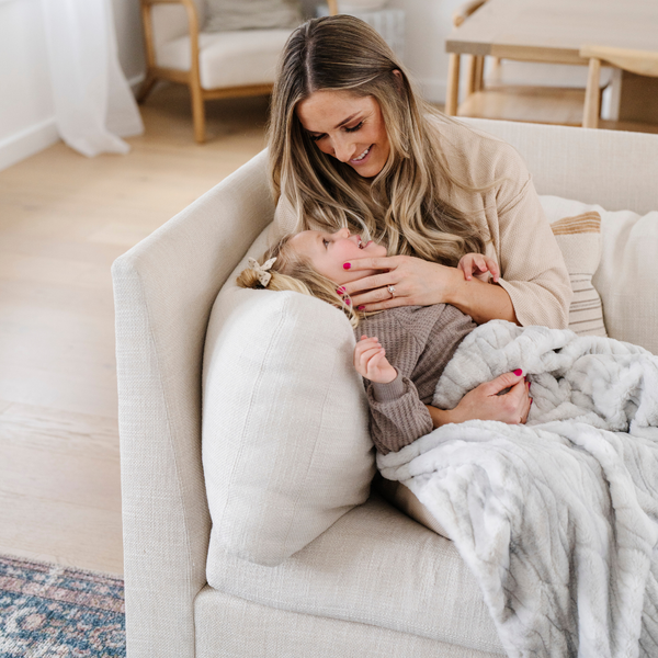 A mother and her daughter sit under a Saranoni blanket, specifically the gray marbled faux fur/minky extra large blanket, highlighting its ultra-soft texture and luxurious design. This plush blanket features a dual-texture blend of silky minky and cozy faux fur, creating the ultimate cozy blanket for home decor. Perfect luxury blanket for lounging or adding style to any space. Soft blanket, cozy blanket—experience unmatched warmth with a Saranoni blanket. 