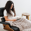 A woman sits in a chair and has a luxurious double-layer Bamboni® blanket by Saranoni, featuring gray leopard print that is white and gray. This ultra-soft blanket is crafted from bamboo rayon and poly microfiber, making it lightweight, breathable, and fluffy. Ideal as a cozy throw blanket, luxury blanket, plush throw, soft blanket, or stylish home accent, this Saranoni blanket brings warmth, comfort, and timeless elegance to any living room or bedroom.