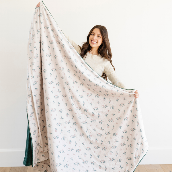 Woman holding a large holiday blanket with a holly berry print and green backing, smiling and showcasing the blanket’s soft and cozy texture. Great for winter home decor and holiday gifting. The soft blanket is a luxury blanket and a Christmas blanket. 