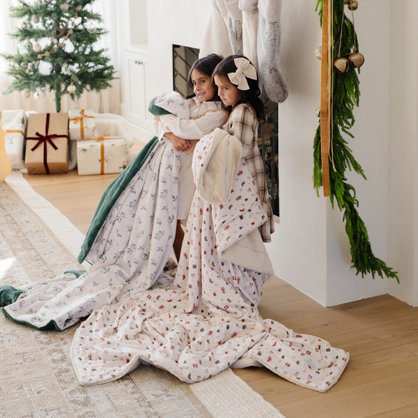 Two young girls bundled in festive holiday blankets by the fireplace, surrounded by Christmas decor. One blanket features a holly berry print, and the other has a holiday-themed pattern with gift boxes. Ideal for holiday gifting and cozy winter moments. The soft blankets are luxury blankets and Christmas blankets. 