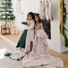 Two girls standing by a fireplace, wrapped in holiday-themed blankets with Christmas decor in the background. The scene includes a decorated tree, wrapped presents, and stockings, perfect for capturing the holiday spirit with cozy throw blankets. One blanket features a holly berry print, and the other has a holiday-themed pattern with gift boxes. Ideal for holiday gifting and cozy winter moments. The soft blankets are luxury blankets and Christmas blankets. 