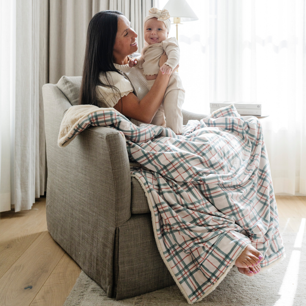 A mother holding her smiling baby, both touching a cozy plaid blanket with red and green accents. Ideal for holiday snuggles and family moments, this plaid throw blanket brings warmth and festive charm to any space. The soft blanket is a luxury blanket and a Christmas blanket. 