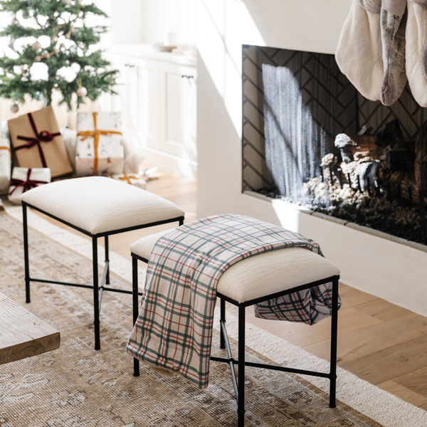 Plaid blanket with red and green stripes draped over a cream ottoman in front of a cozy fireplace with wrapped holiday gifts and a decorated Christmas tree. Perfect for holiday decor and adding warmth to your living room during winter. The soft blanket is a luxury blanket and a Christmas blanket. 