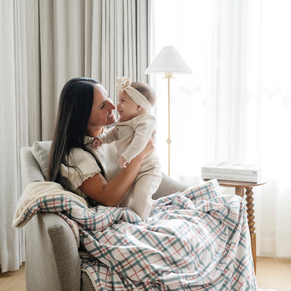 A mother holding her smiling baby, both touching a cozy plaid blanket with red and green accents. Ideal for holiday snuggles and family moments, this plaid throw blanket brings warmth and festive charm to any space. The soft blanket is a luxury blanket and a Christmas blanket. 