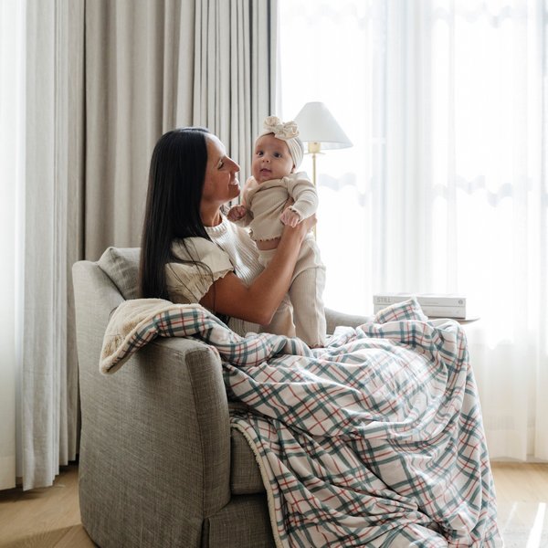 A mother holding her smiling baby, both touching a cozy plaid blanket with red and green accents. Ideal for holiday snuggles and family moments, this plaid throw blanket brings warmth and festive charm to any space. The soft blanket is a luxury blanket and a Christmas blanket. 