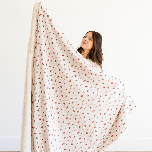 Woman holding up a holiday blanket with a festive pattern featuring gift boxes, stockings, Christmas trees, and other holiday illustrations. Ideal for cozy winter decor and holiday gifts that bring warmth and joy. The soft blanket is a luxury blanket and a Christmas blanket. 