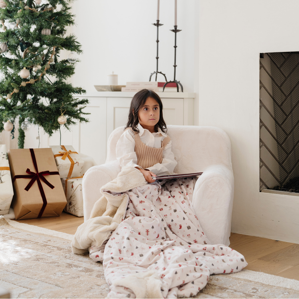 Young girl sitting on a cozy armchair next to a decorated Christmas tree, wrapped in a holiday blanket with festive gift boxes, stockings, and more holiday illustrations. Perfect for holiday decor and creating warm, seasonal family moments. The soft blanket is a luxury blanket and a Christmas blanket. 