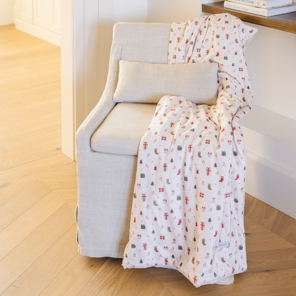 Holiday blanket with a charming pattern of gift boxes, Christmas trees, and stockings draped over a beige armchair, adding a festive touch to home decor. Perfect for seasonal warmth and holiday styling. The soft blanket is a luxury blanket and a Christmas blanket. 